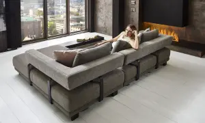 Moderne Wohnlandschaft mit grauem Stoffbezug, die als **Sofa als Raumteiler Ideen** dient. Eine Frau entspannt sich darauf, im Hintergrund ein Kaminfeuer.