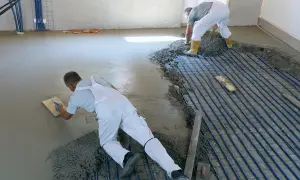 Zwei Männer machen Estrich selber, einer glättet den frischen Beton, der andere verteilt ihn über Heizungsrohren.