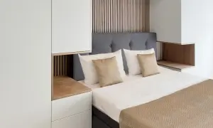 Ein gemütliches kleines Schlafzimmer einrichten mit einem Bett, grauem Kopfteil, weißen und goldenen Kissen, Holzakzenten und weißen Schränken.