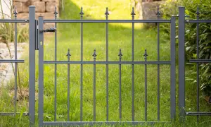 Graues Metallgartentor mit Griff und Schloss, bereit zum **Gartentor einbetonieren**. Dahinter grüner Rasen und eine Steinmauer.