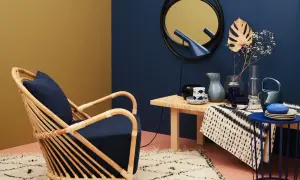 Ein Rattansessel mit dunkelblauem Kissen steht auf einem Teppich. Welche Farbe passt zu Gold? Hier: ein goldener Spiegel und eine gelbliche Wand.