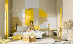 Eine Frau entspannt sich in einem hellen Wohnzimmer mit geometrischen Wandmustern. Die Frage, welche Farbe passt zu Gelb, wird hier mit Beige und Grau beantwortet.