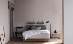 Ein helles, skandinavisches Schlafzimmer mit einem Holzbett, grauen Kissen und einer Stehlampe. Zwei gerollte Decken hängen an der Wand.