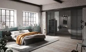 Ein gemütliches bett unter fenster mit Blick auf die Stadt. Daneben ein Nachttisch und eine Pflanze. Im Hintergrund ein offenes Badezimmer.