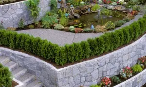 Eine malerische Gartentreppe führt an einer rustikalen steinmauer entlang, vorbei an einem kleinen Teich mit Wasserfall und üppiger Bepflanzung.