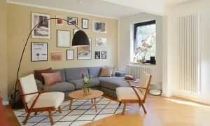 Gemütliches Wohnzimmer mit grauem Sofa, zwei Sesseln und einem Couchtisch. Die gelbe Wand ist mit vielen Bildern dekoriert, die den Rand des Raumes betonen.
