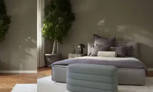Ein stilvolles Schlafzimmer in gedeckten schlafzimmer farben mit zwei großen Pflanzen, einem Bett mit vielen Kissen und einem grauen Hocker.