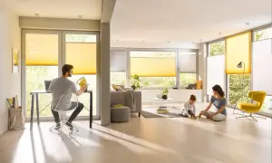 Familie genießt das Leben in einem hellen Wohnzimmer mit gelben Plissees, die das Fenster gestalten. Ein Mann arbeitet am Laptop, während Mutter und Kind spielen.