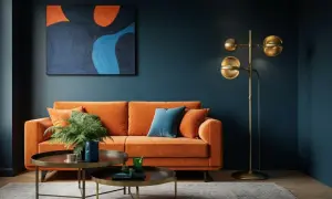 Ein orangefarbenes Sofa mit Kissen und einer Pflanze steht vor einer dunkelblauen Wand. Ein abstraktes Gemälde mit warmen Farben hängt darüber. Ein goldener Stehlampen und zwei Couchtische vervollständigen das Bild.
