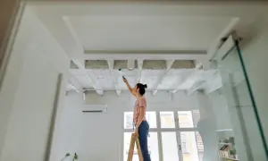 Frau streicht Decke mit Farbroller. Sie steht auf Leiter, um ihre Wohnung zu renovieren.