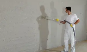 Mann verputzt eine Wand mit Spritzputz. So wird eine Wand verputzt: Gleichmäßige Bewegung der Spritzpistole.
