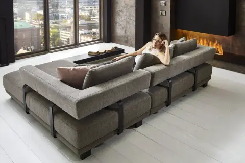 Moderne Wohnlandschaft mit grauem Stoffbezug, die als **Sofa als Raumteiler Ideen** dient. Eine Frau entspannt sich darauf, im Hintergrund ein Kaminfeuer.