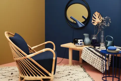Ein Rattansessel mit dunkelblauem Kissen steht auf einem Teppich. Welche Farbe passt zu Gold? Hier: ein goldener Spiegel und eine gelbliche Wand.
