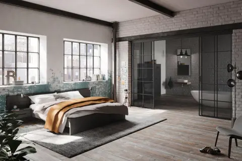Ein gemütliches bett unter fenster mit Blick auf die Stadt. Daneben ein Nachttisch und eine Pflanze. Im Hintergrund ein offenes Badezimmer.