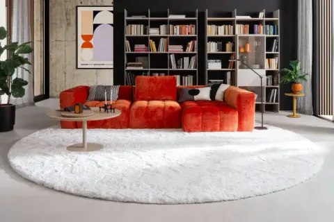 Ein flauschiger, weißer Teppich liegt unter dem auffälligen, orangefarbenen Sofa und dem runden Couchtisch.