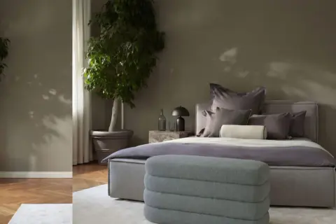Ein stilvolles Schlafzimmer in gedeckten schlafzimmer farben mit zwei großen Pflanzen, einem Bett mit vielen Kissen und einem grauen Hocker.