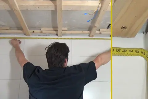 Mann misst die Decke für das Anbringen von Deckenpaneelen. Holzlatten sind bereits montiert.