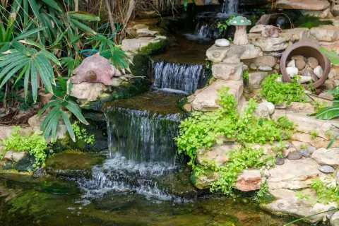 Ein kleiner Wasserfall plätschert in einen Teich, der mit Steinen und Pflanzen gestaltet ist. Eine Schildkrötenfigur wacht über das Wasser.