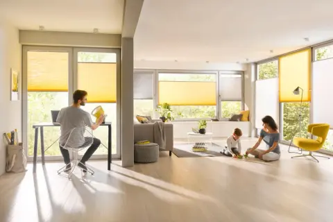 Familie genießt das Leben in einem hellen Wohnzimmer mit gelben Plissees, die das Fenster gestalten. Ein Mann arbeitet am Laptop, während Mutter und Kind spielen.