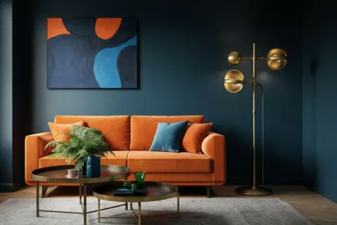 Ein orangefarbenes Sofa mit Kissen und einer Pflanze steht vor einer dunkelblauen Wand. Ein abstraktes Gemälde mit warmen Farben hängt darüber. Ein goldener Stehlampen und zwei Couchtische vervollständigen das Bild.