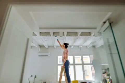 Frau streicht Decke mit Farbroller. Sie steht auf Leiter, um ihre Wohnung zu renovieren.