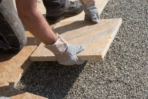 Arbeiter verlegt sorgfältig eine helle Terrassenplatte auf Kies.