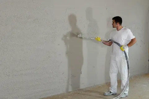 Mann verputzt eine Wand mit Spritzputz. So wird eine Wand verputzt: Gleichmäßige Bewegung der Spritzpistole.