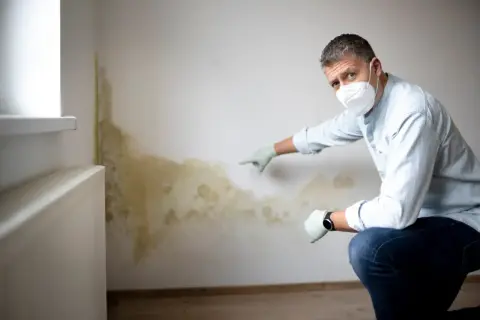 Mann mit Maske und Handschuhen zeigt auf deutlichen **Schimmel an der Wand**, der sich neben einem Heizkörper ausgebreitet hat.