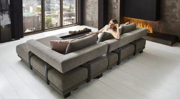 Moderne Wohnlandschaft mit grauem Stoffbezug, die als **Sofa als Raumteiler Ideen** dient. Eine Frau entspannt sich darauf, im Hintergrund ein Kaminfeuer.