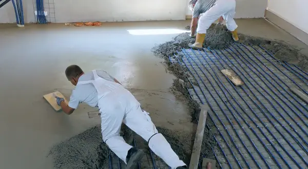 Zwei Männer machen Estrich selber, einer glättet den frischen Beton, der andere verteilt ihn über Heizungsrohren.
