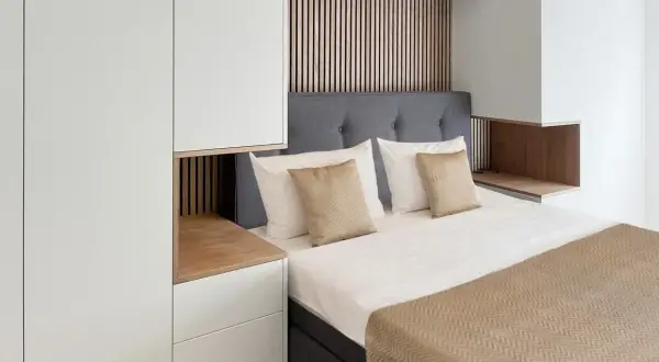 Ein gemütliches kleines Schlafzimmer einrichten mit einem Bett, grauem Kopfteil, weißen und goldenen Kissen, Holzakzenten und weißen Schränken.