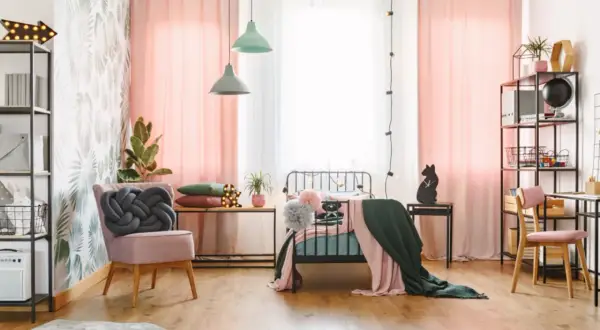 Ein stilvolles Jugendzimmer einrichten: Rosa Vorhänge, ein Bett mit Decken, ein Sessel mit Knoten-Kissen und ein Schreibtisch mit Regal.