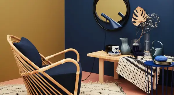 Ein Rattansessel mit dunkelblauem Kissen steht auf einem Teppich. Welche Farbe passt zu Gold? Hier: ein goldener Spiegel und eine gelbliche Wand.