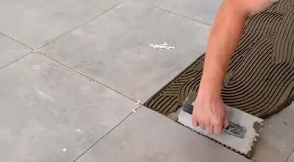 Hand spreadet Mörtel mit einer Kelle, um Fliesen zu verlegen. Kleine weiße Abstandshalter sorgen für gleichmäßige Fugen.