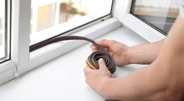 Hände halten eine Rolle Dichtungsband bereit, um ein Fenster abzudichten.