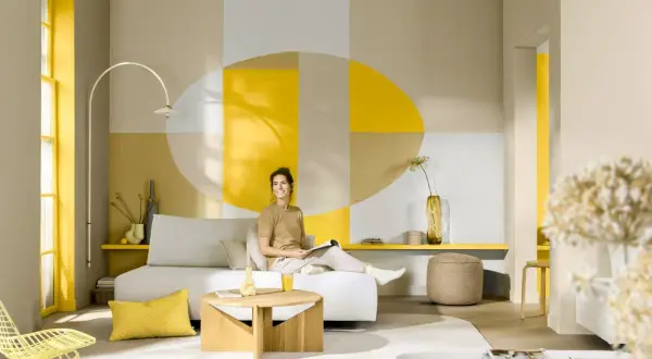 Eine Frau entspannt sich in einem hellen Wohnzimmer mit geometrischen Wandmustern. Die Frage, welche Farbe passt zu Gelb, wird hier mit Beige und Grau beantwortet.