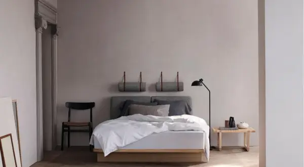 Ein helles, skandinavisches Schlafzimmer mit einem Holzbett, grauen Kissen und einer Stehlampe. Zwei gerollte Decken hängen an der Wand.