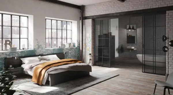Ein gemütliches bett unter fenster mit Blick auf die Stadt. Daneben ein Nachttisch und eine Pflanze. Im Hintergrund ein offenes Badezimmer.