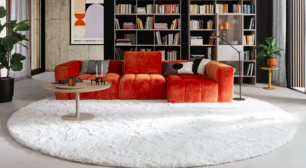 Ein flauschiger, weißer Teppich liegt unter dem auffälligen, orangefarbenen Sofa und dem runden Couchtisch.