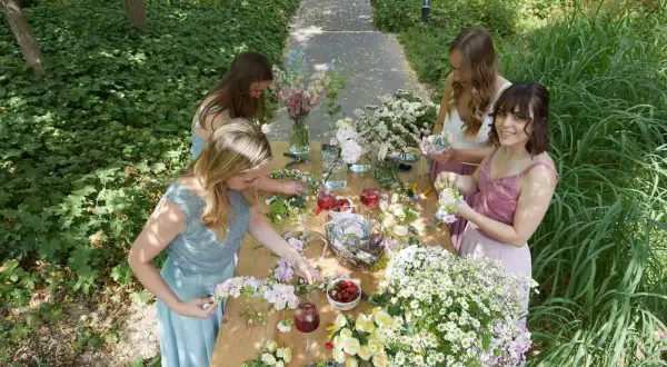 Vier Frauen binden im Freien einen Blumenkranz. Sie sitzen an einem Holztisch voller Blumen, Werkzeuge und Getränke.