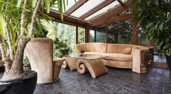 Gemütliches Ambiente im Wintergarten: Einladende Couch, geschwungener Glastisch und üppige Pflanzen laden zum Entspannen ein. Perfekt, um den Wintergarten einzurichten.