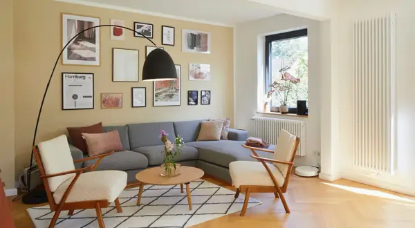 Gemütliches Wohnzimmer mit grauem Sofa, zwei Sesseln und einem Couchtisch. Die gelbe Wand ist mit vielen Bildern dekoriert, die den Rand des Raumes betonen.