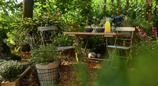 Gemütliche Gartenecke mit Tisch und Stühlen, umgeben von üppigem Grün. Rindenmulch bedeckt den Boden und bietet tolle Beet-Ideen für naturnahe Gestaltung.