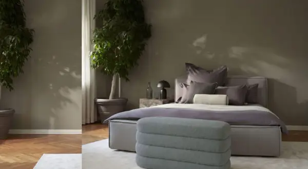 Ein stilvolles Schlafzimmer in gedeckten schlafzimmer farben mit zwei großen Pflanzen, einem Bett mit vielen Kissen und einem grauen Hocker.