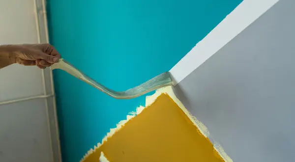 Hand zieht Klebeband ab, um eine gerade Kante zwischen türkisfarbener und grauer Wand zu schaffen. So gelingt das **richtig streichen**.