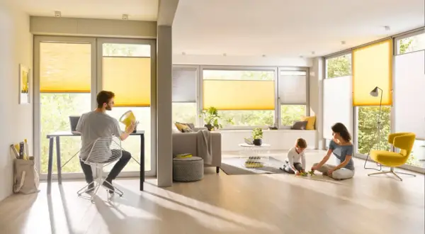Familie genießt das Leben in einem hellen Wohnzimmer mit gelben Plissees, die das Fenster gestalten. Ein Mann arbeitet am Laptop, während Mutter und Kind spielen.