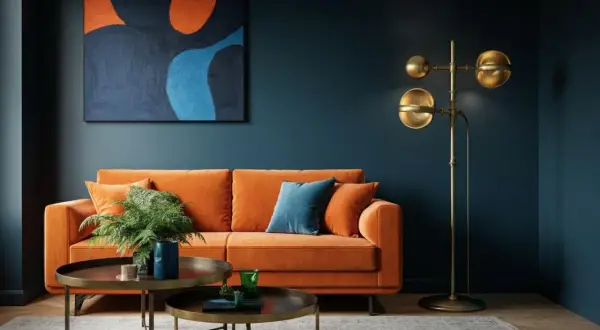 Ein orangefarbenes Sofa mit Kissen und einer Pflanze steht vor einer dunkelblauen Wand. Ein abstraktes Gemälde mit warmen Farben hängt darüber. Ein goldener Stehlampen und zwei Couchtische vervollständigen das Bild.