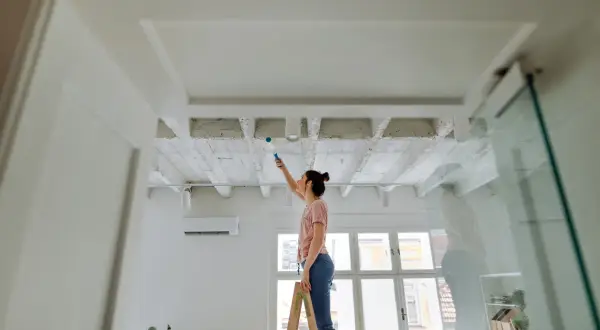 Frau streicht Decke mit Farbroller. Sie steht auf Leiter, um ihre Wohnung zu renovieren.