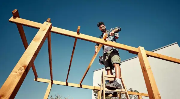 Mann auf Leiter schraubt Holzkonstruktion für ein Terrassendach selber bauen.