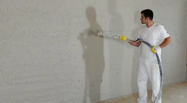 Mann verputzt eine Wand mit Spritzputz. So wird eine Wand verputzt: Gleichmäßige Bewegung der Spritzpistole.